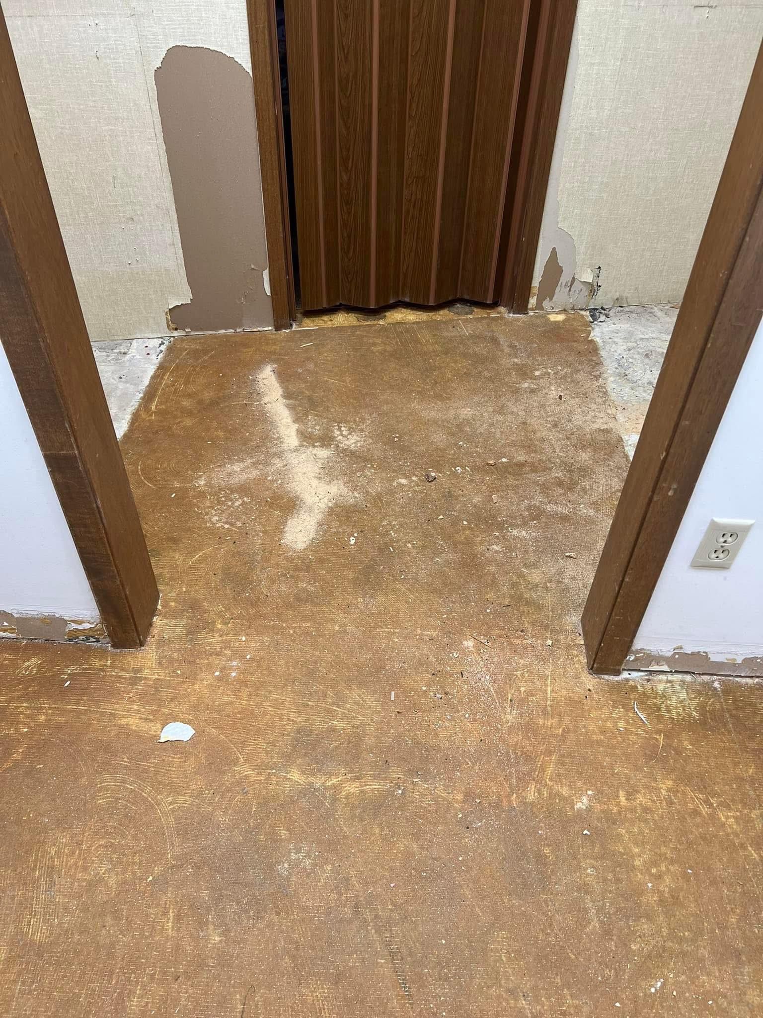 Doorway with damaged flooring, brown trim, and wall. White electrical outlet on right.