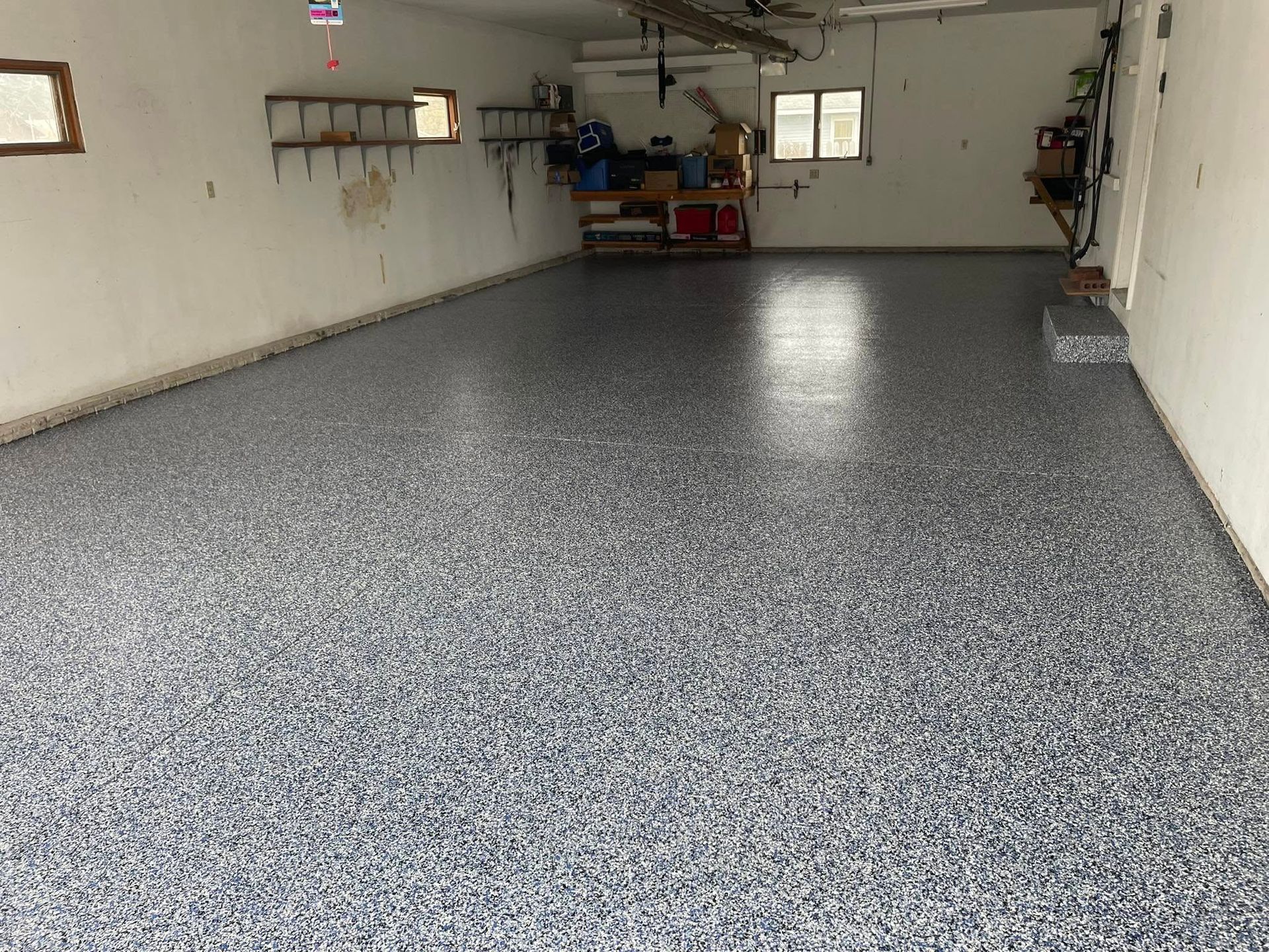 A garage interior with a gray, speckled epoxy floor. White walls, empty shelves, and two small windows are visible.