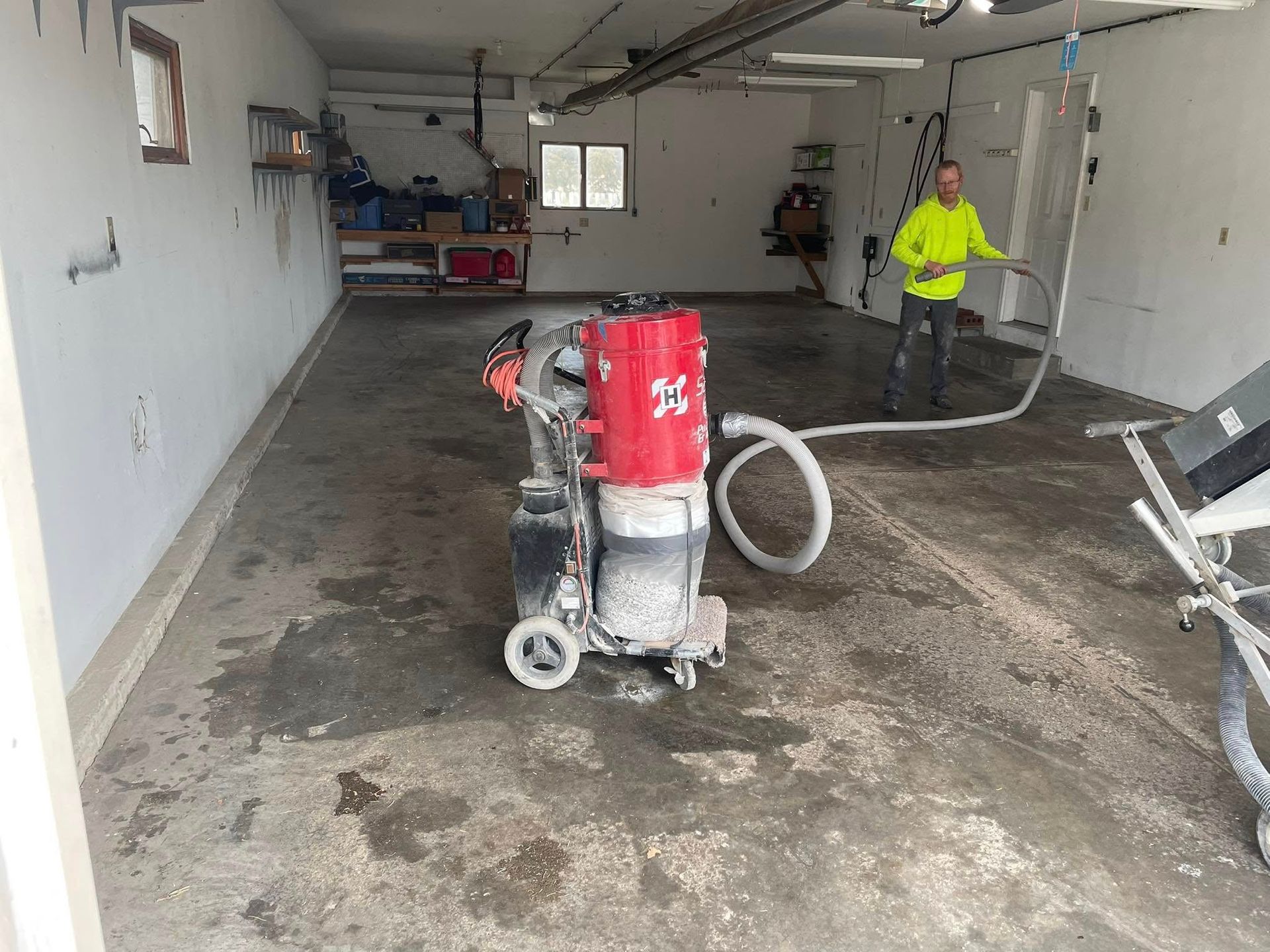 Man grinding concrete floor in a garage with large dust collection machine, white walls.