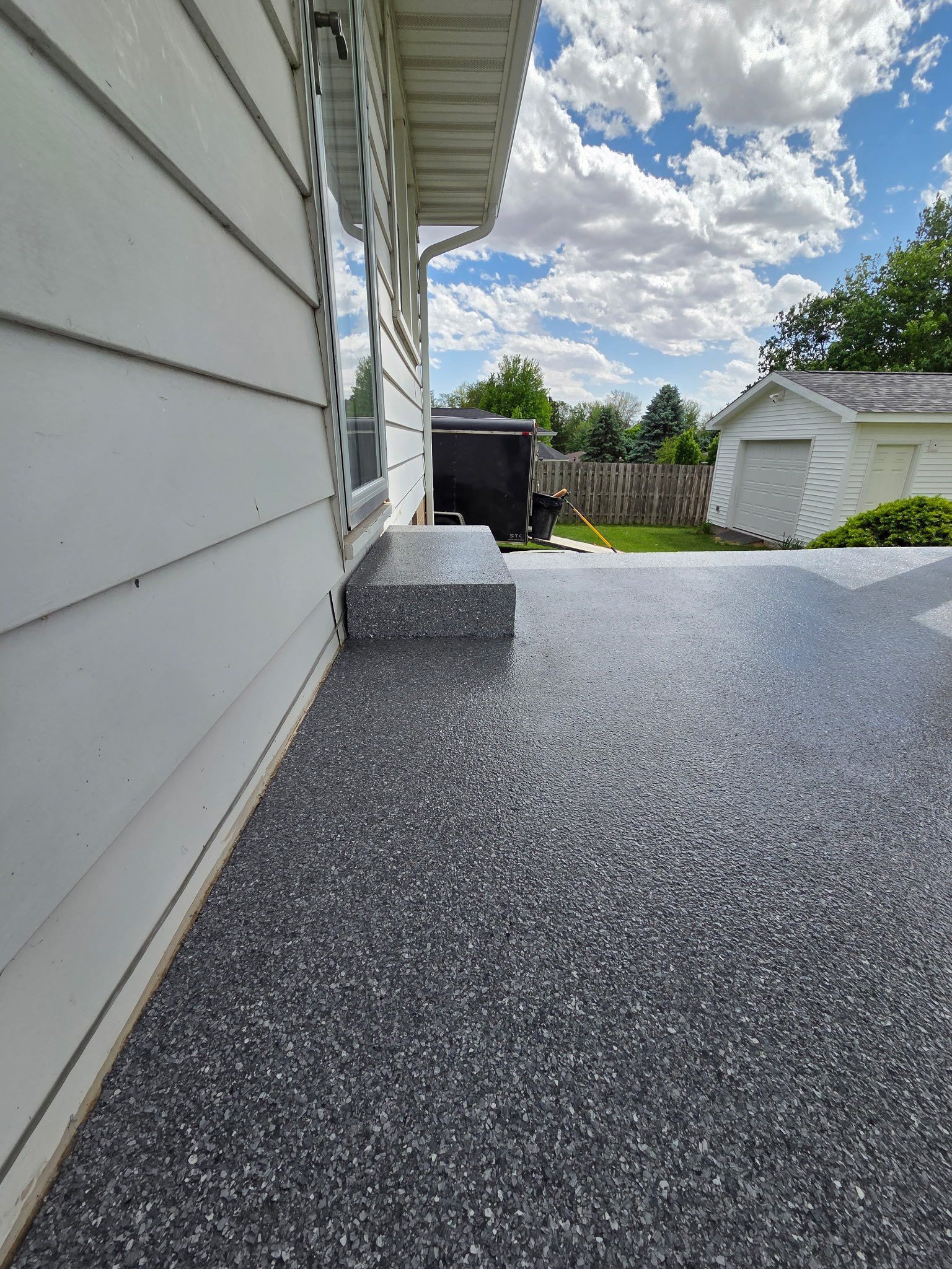 Exterior patio with gray speckled flooring, step, and white siding. Sunny, blue sky with clouds.