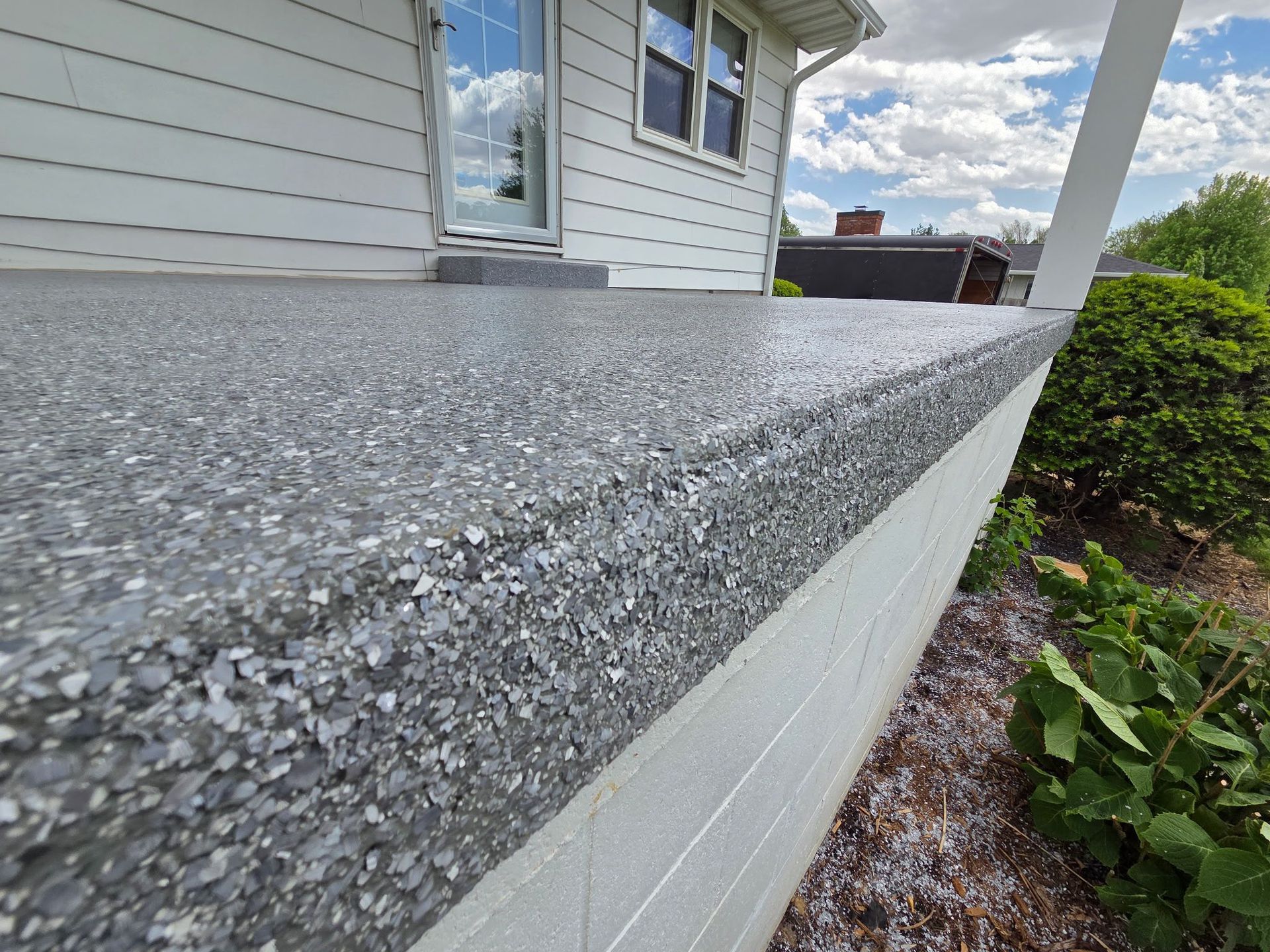 Gray, speckled concrete porch with a white house, window, and shrubbery in the background.