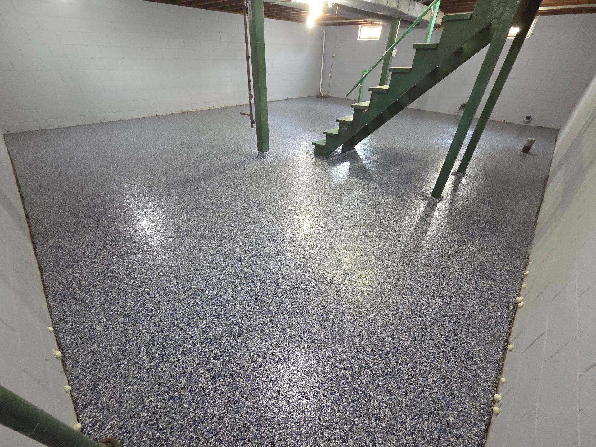 Basement with a speckled epoxy floor, green stairs, and support beams. The room is mostly empty with white walls.