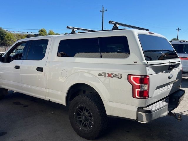 A white Ford F-150 pickup truck with a camper shell and roof rack, parked outdoors on a sunny day.