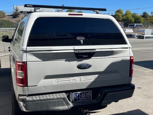 Rear view of a white Ford F-150 pickup truck with a camper shell and roof rack, parked outdoors on a sunny day.