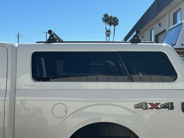 A white pickup truck camper shell featuring a roof rack with two black crossbars, parked outdoors on a sunny day.