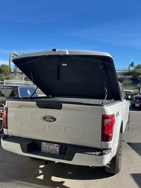 A white Ford pickup truck parked outdoors with its black tonneau bed cover lifted open.
