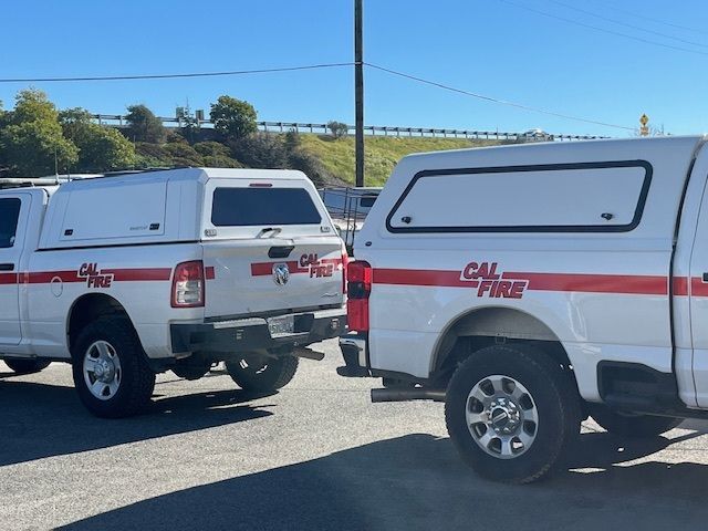 Two white Cal Fire pickup trucks with red branding parked side-by-side on an asphalt lot under a clear blue sky.