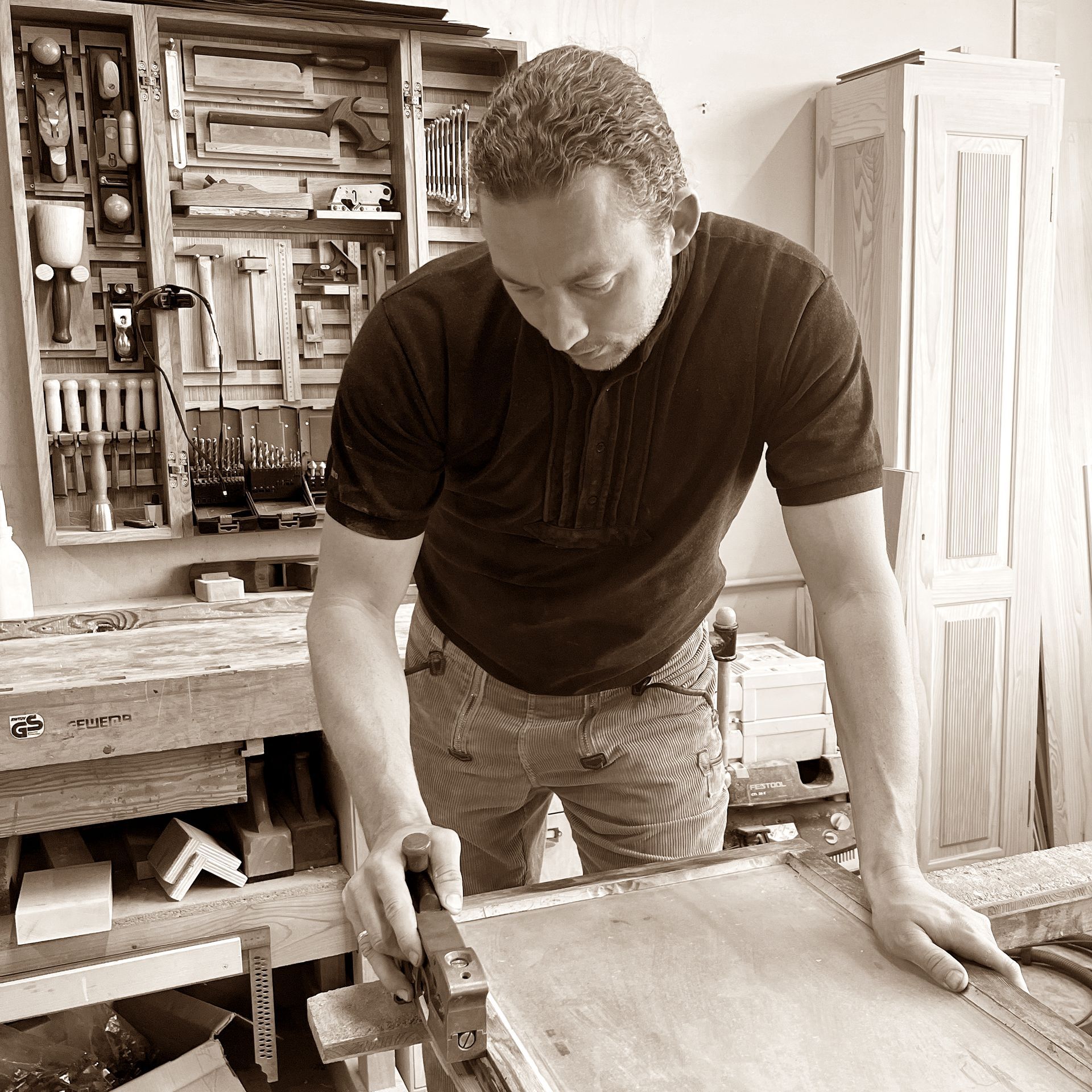 Ein Tischler arbeitet an einer Werkbank und hobelt eine Holzplatte. Er befindet sich in einer Werkstatt mit Werkzeugen an der Wand und Holzmöbeln.