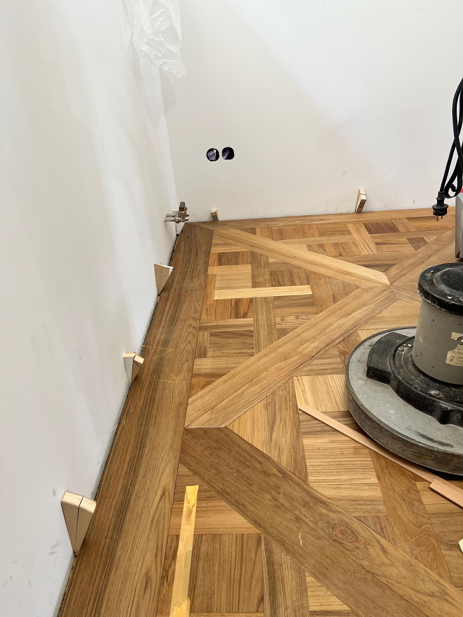 Parkettboden wird mit einer Maschine neben einer weißen Wand mit Steckdosen geschliffen; Holzabstandshalter säumen die Wand.