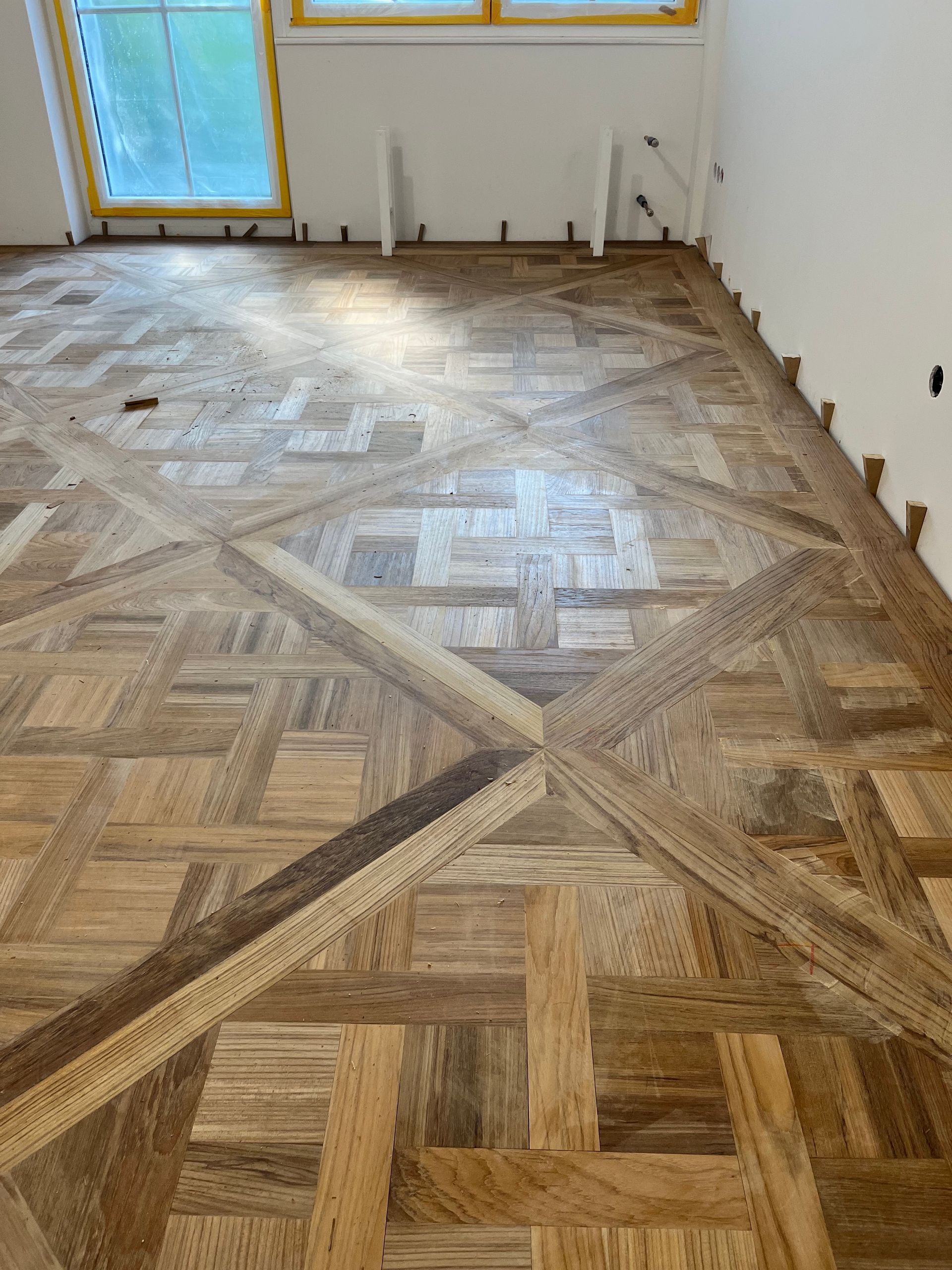 Aufwendige Holzbodenverlegung mit einem sich wiederholenden geometrischen Muster; helle und dunkle Holztöne. Der Boden befindet sich in der Nähe eines Fensters.