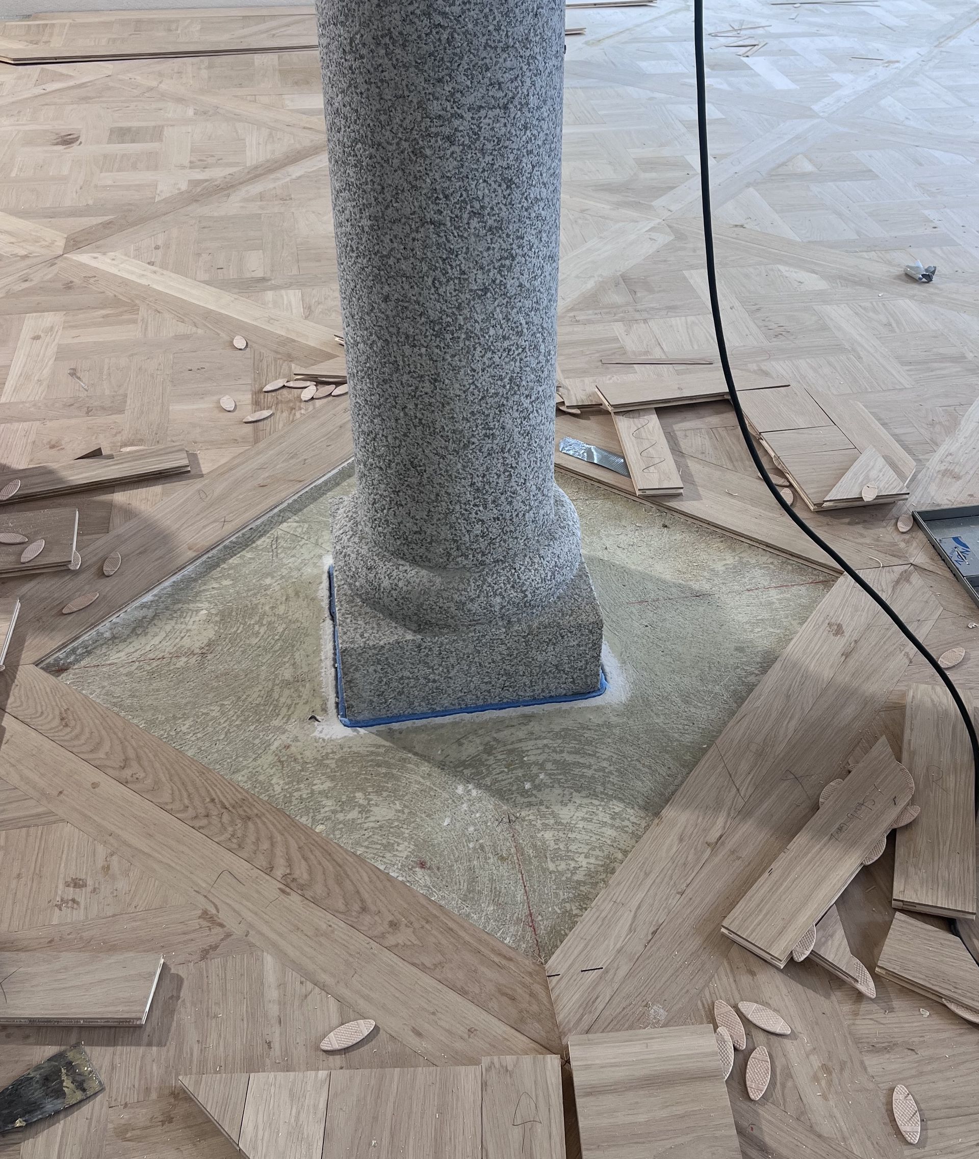 Holzbodenverlegung rund um eine graue Säule. Der Boden weist komplizierte geometrische Muster und verstreute Holzstücke auf.