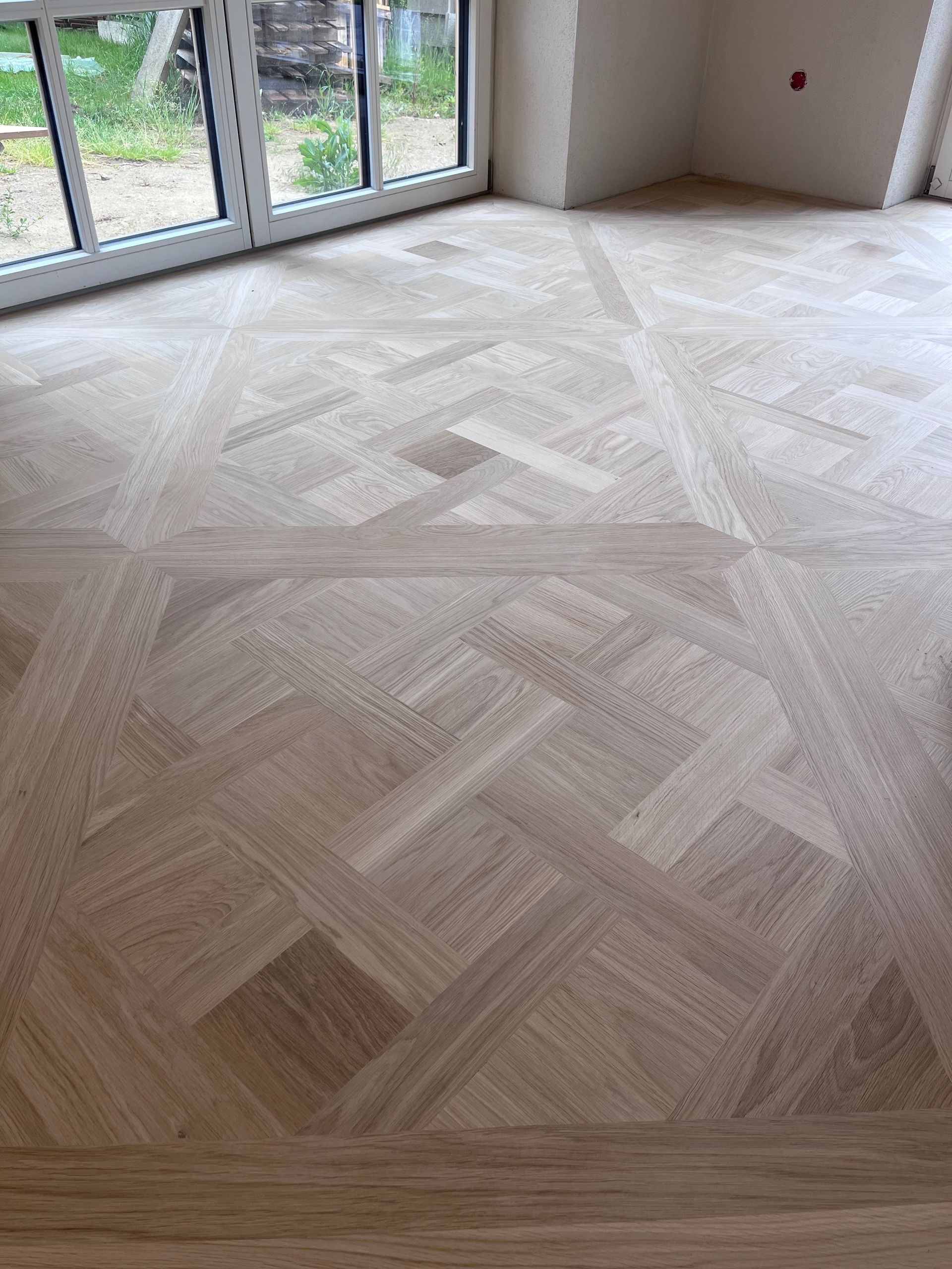 Heller Holzparkettboden mit geometrischen Mustern in einem Raum mit großem Fenster.