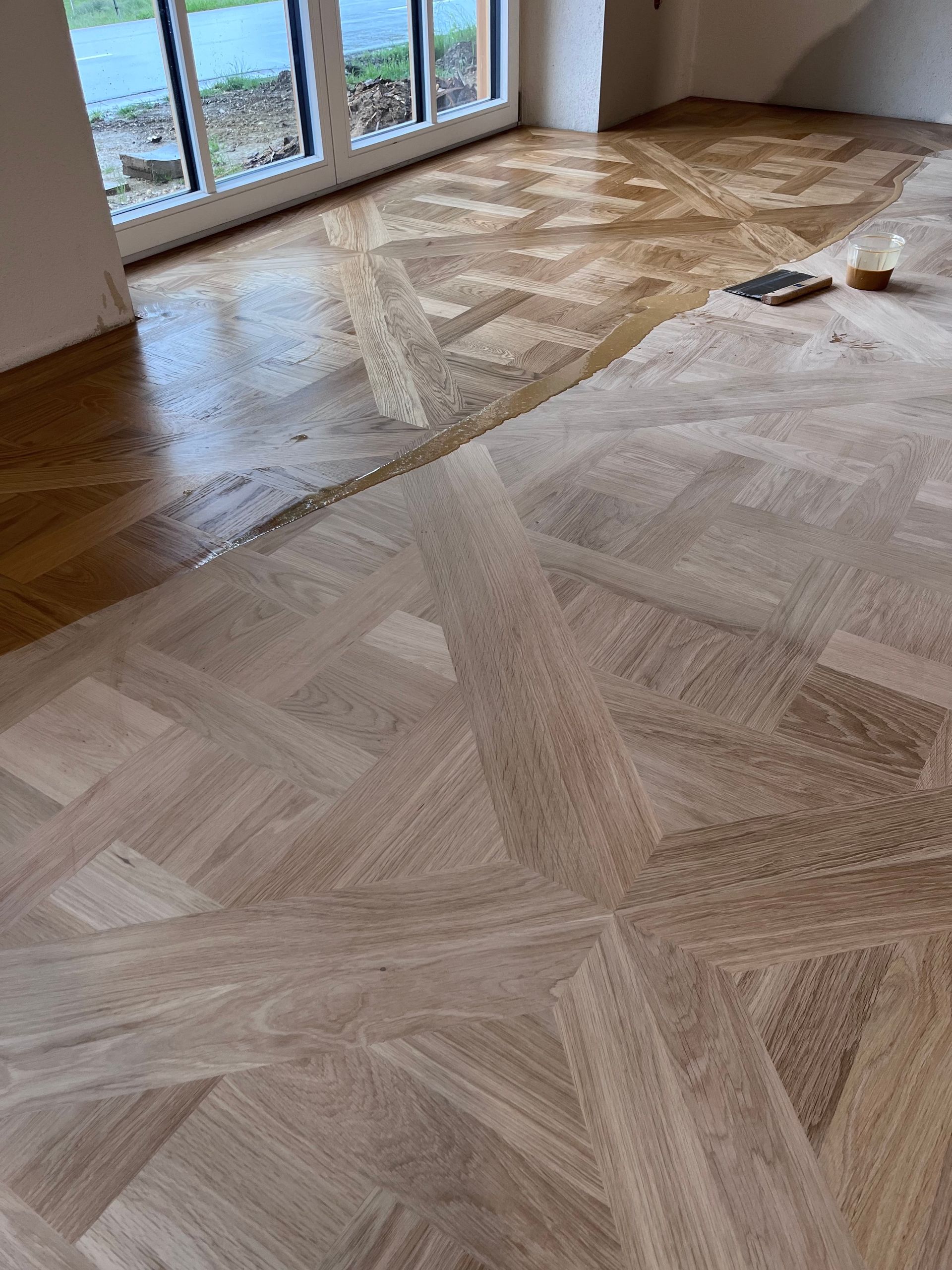 Holzparkettboden in geometrischem Muster, wobei eine Hälfte versiegelt und die andere Hälfte unbehandelt ist. Im Hintergrund ist ein Fenster zu sehen.