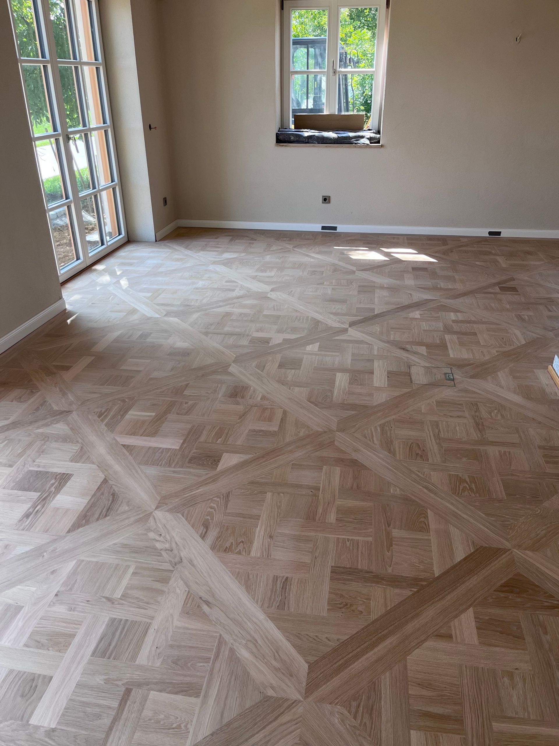 Holzparkettboden in einem Raum mit natürlichem Licht durch Fenster und eine Tür. Der Boden hat ein geometrisches Muster in hellbraunen Tönen.