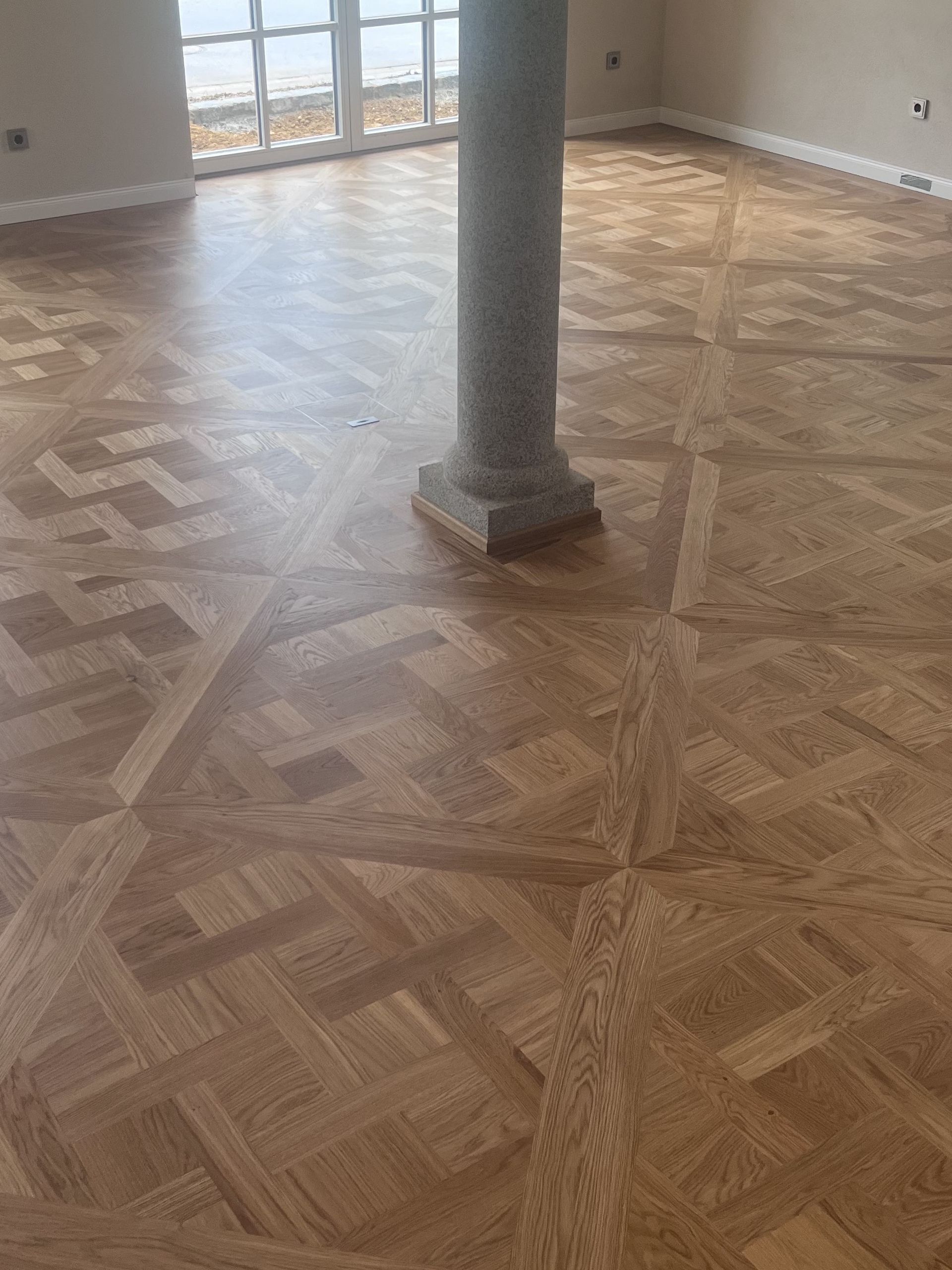 Hellbrauner Parkettboden mit gemustertem Design um eine graue Steinsäule in einem Raum mit hellem Fenster.
