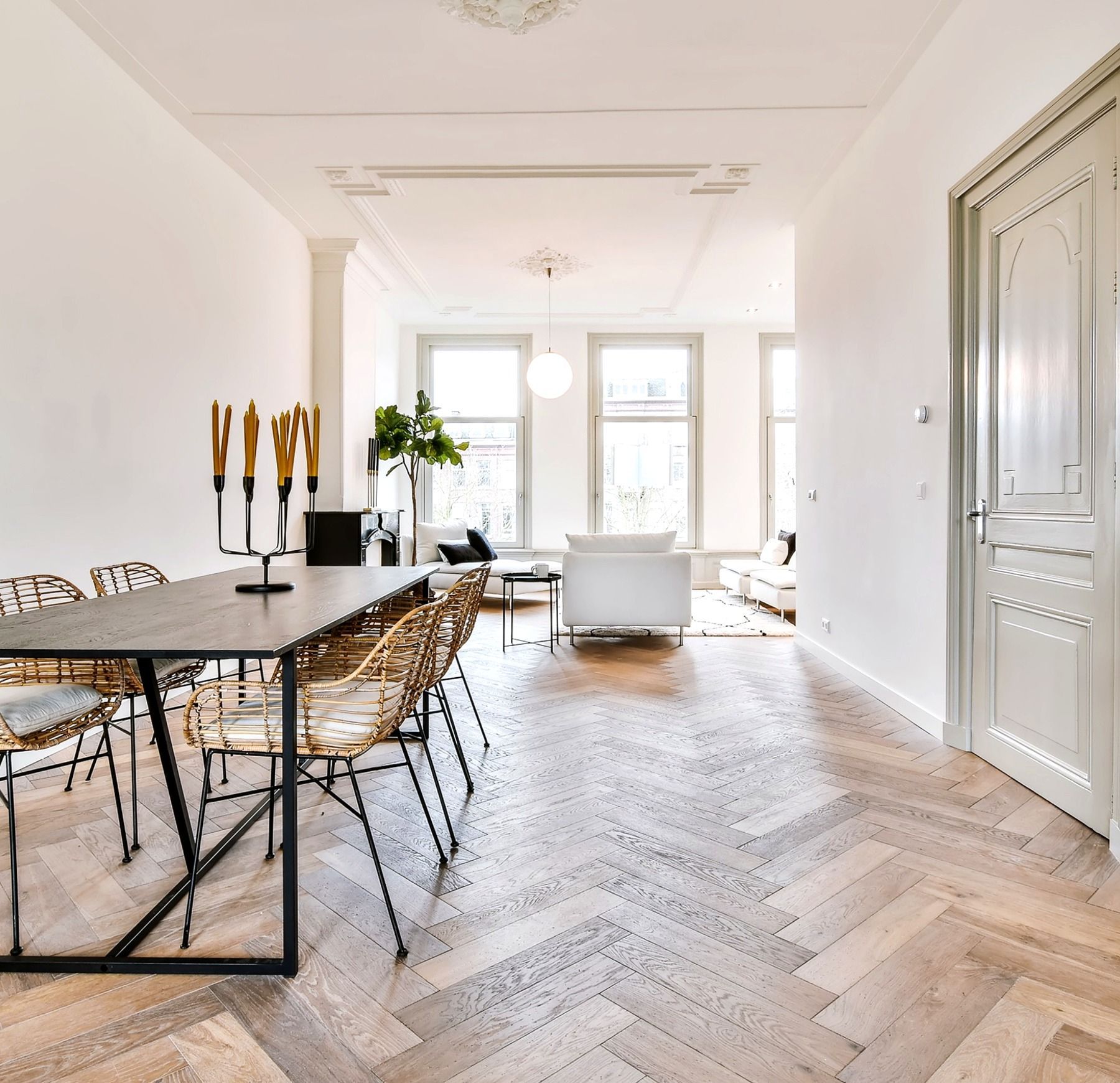A dining table with chairs in the foreground, opening into a bright, modern living room with herringbone wood floors.
