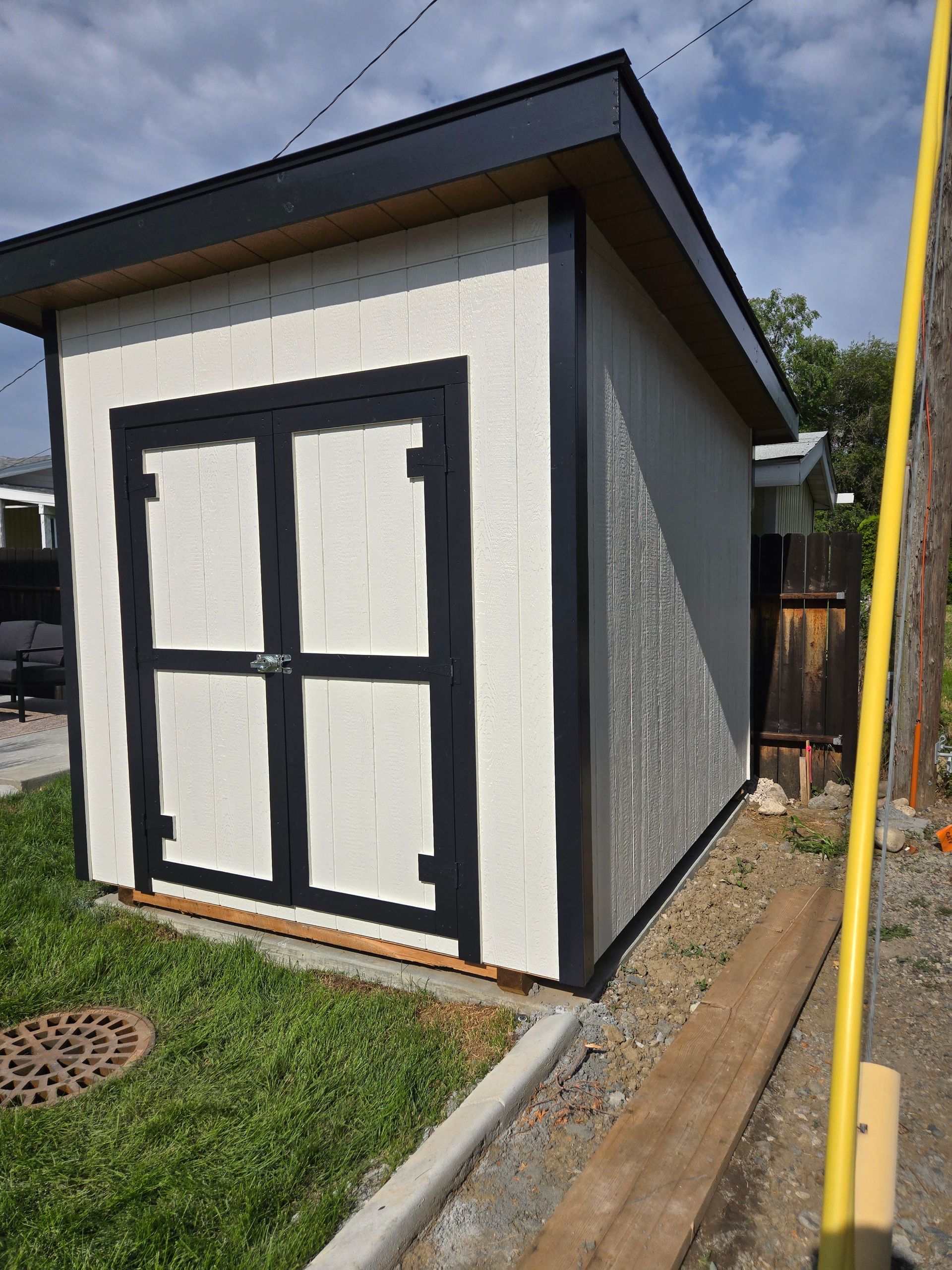 White shed with black trim and doors, located outdoors next to a grassy area and a gravel path.