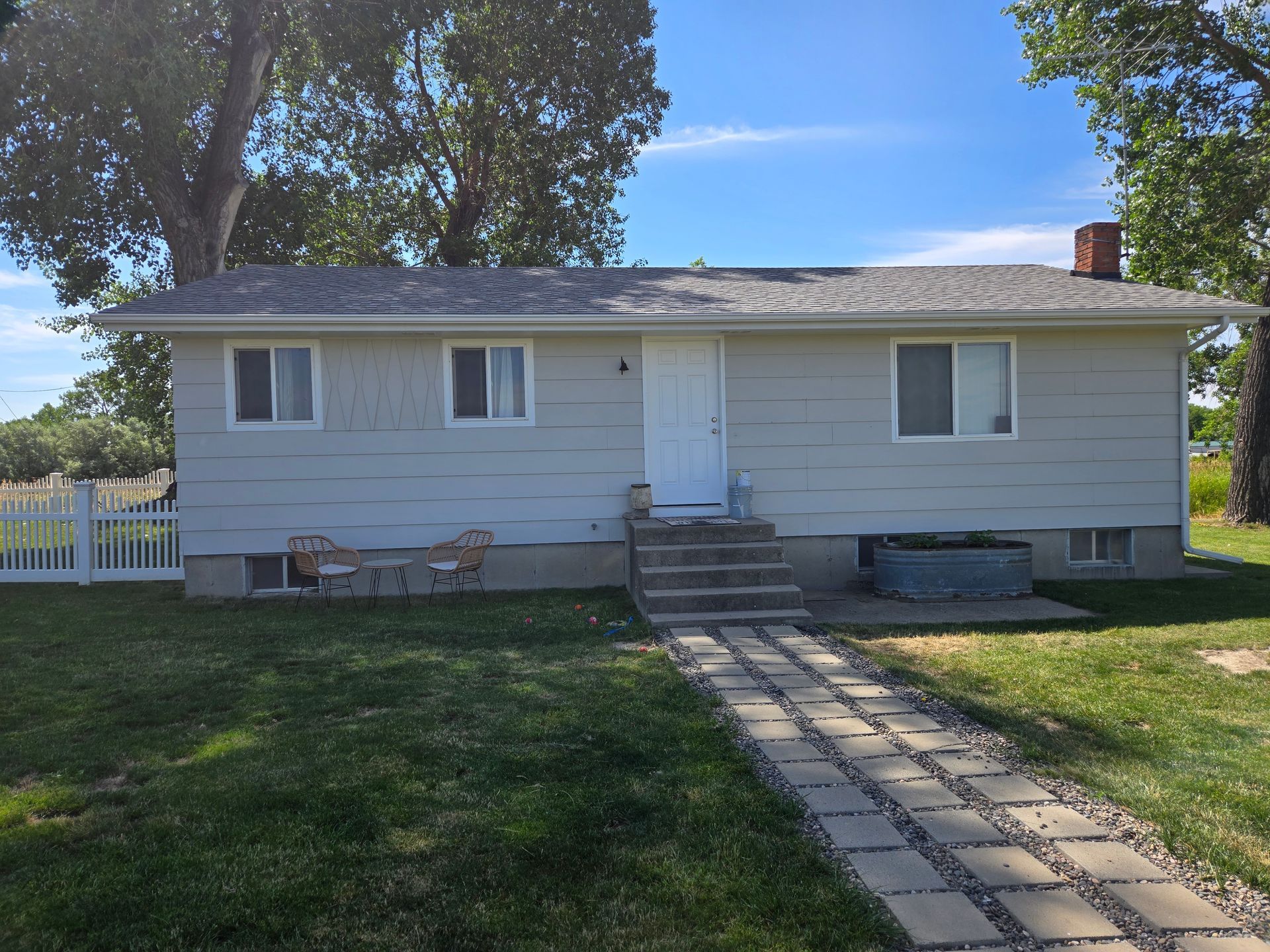 A light gray ranch-style house with a brick walkway and white picket fence on a sunny day.