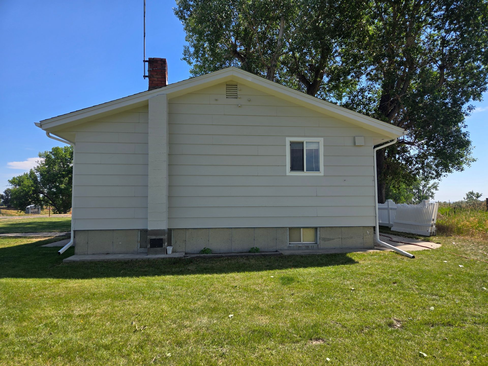 Side view of a light-colored house with a chimney, a small window, and green grass in a rural setting.