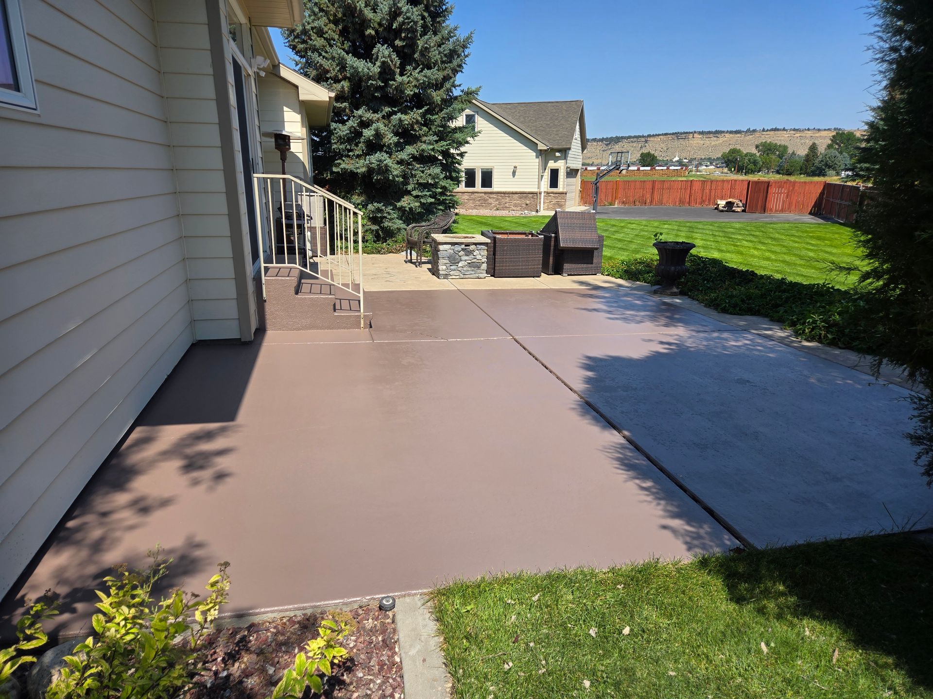 Brown painted concrete patio next to a house with outdoor furniture and a green lawn on a sunny day.