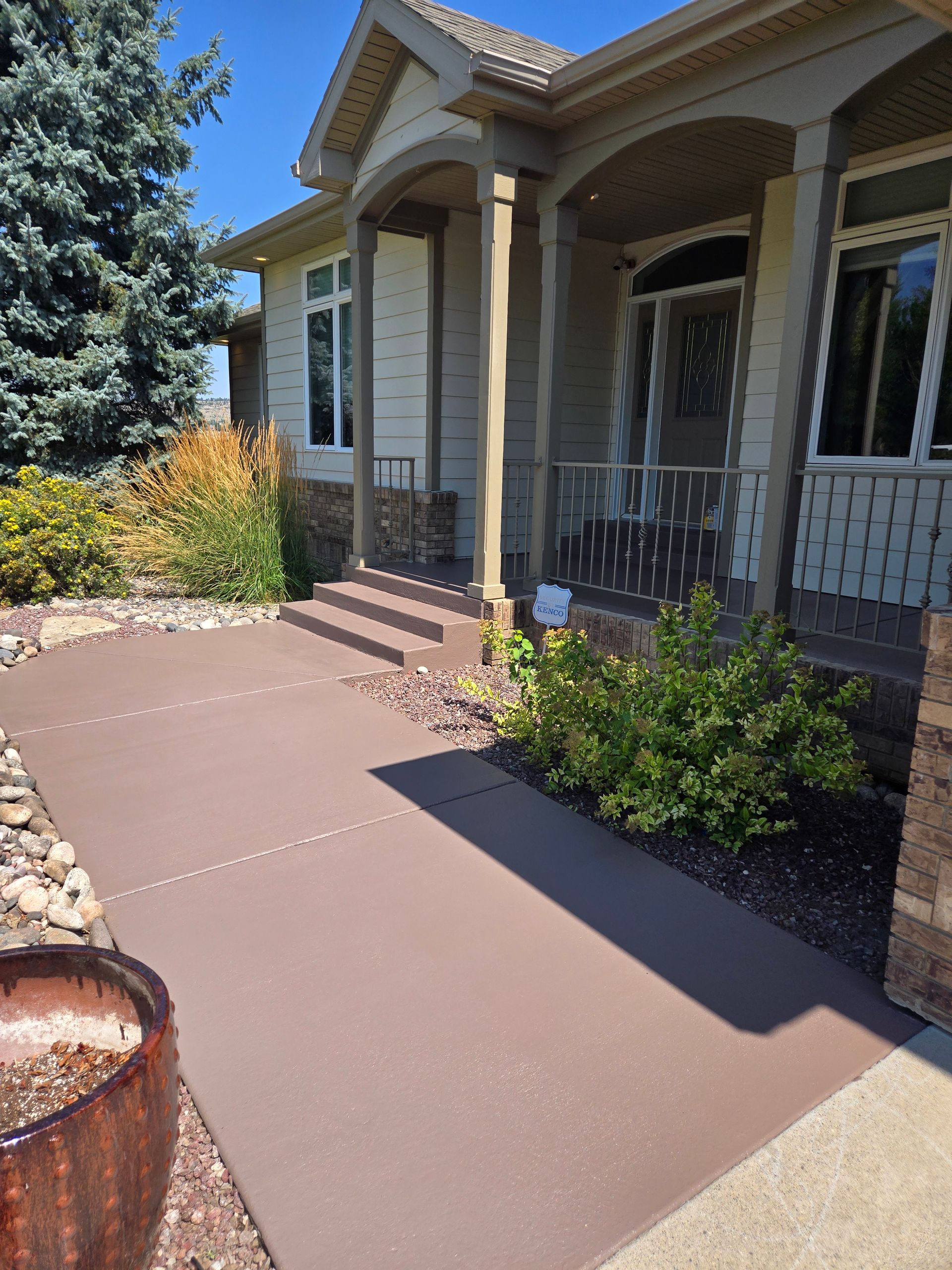 Brown concrete walkway leading to the entrance of a house with beige siding and a porch.