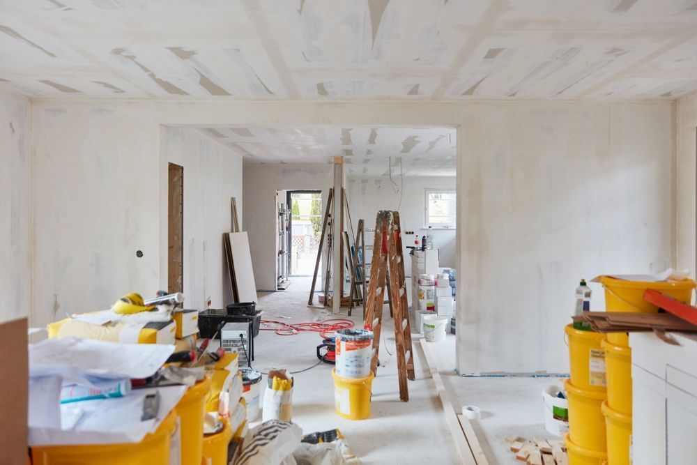 Interior of a room under construction, with white walls and ceiling. Ladders, tools, and materials scattered.