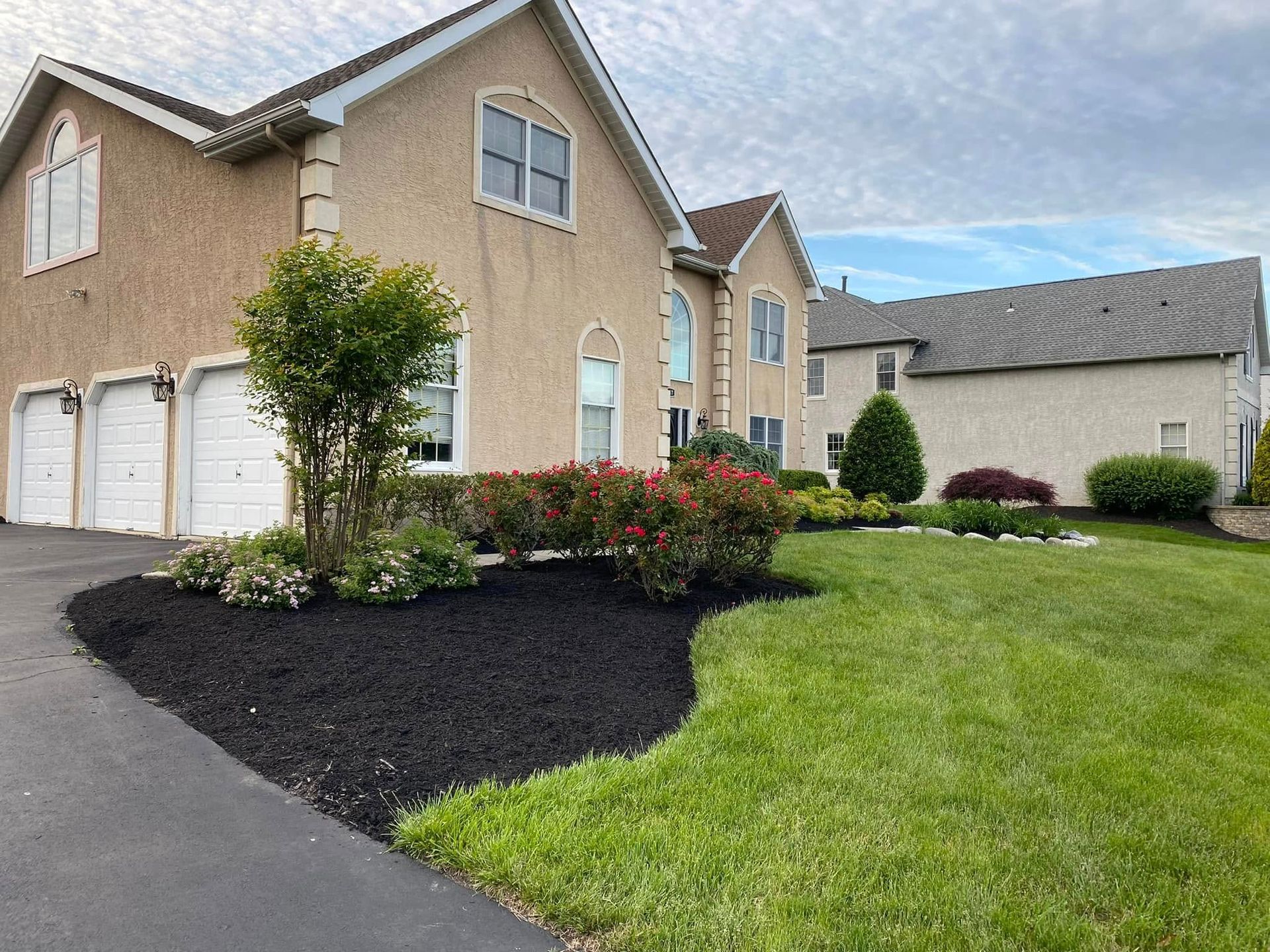 Two-story beige house with three-car garage, manicured lawn, black mulch, and landscaping under a cloudy sky.