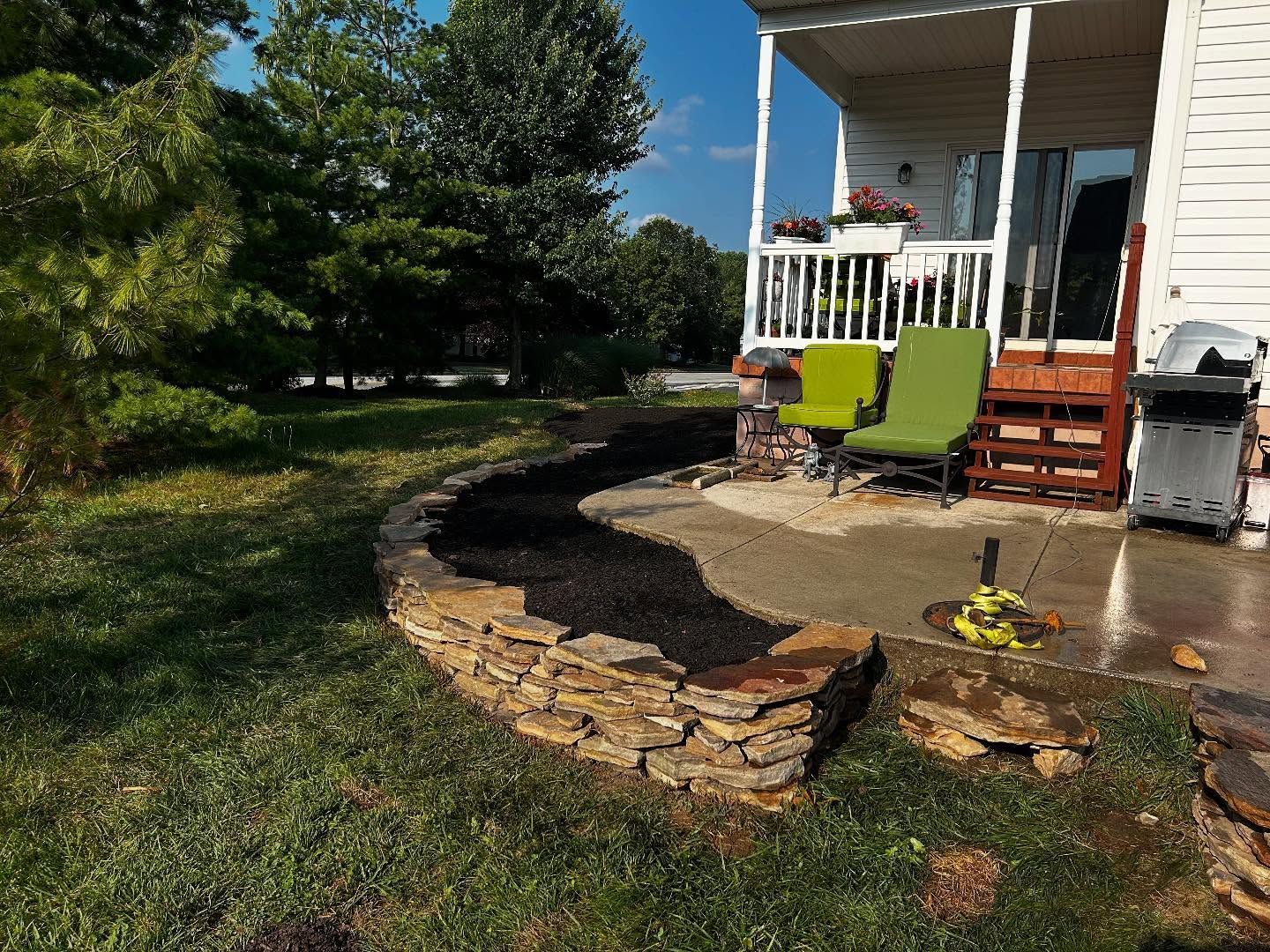 Backyard with a stone retaining wall filled with mulch. Patio with green chairs, grill.
