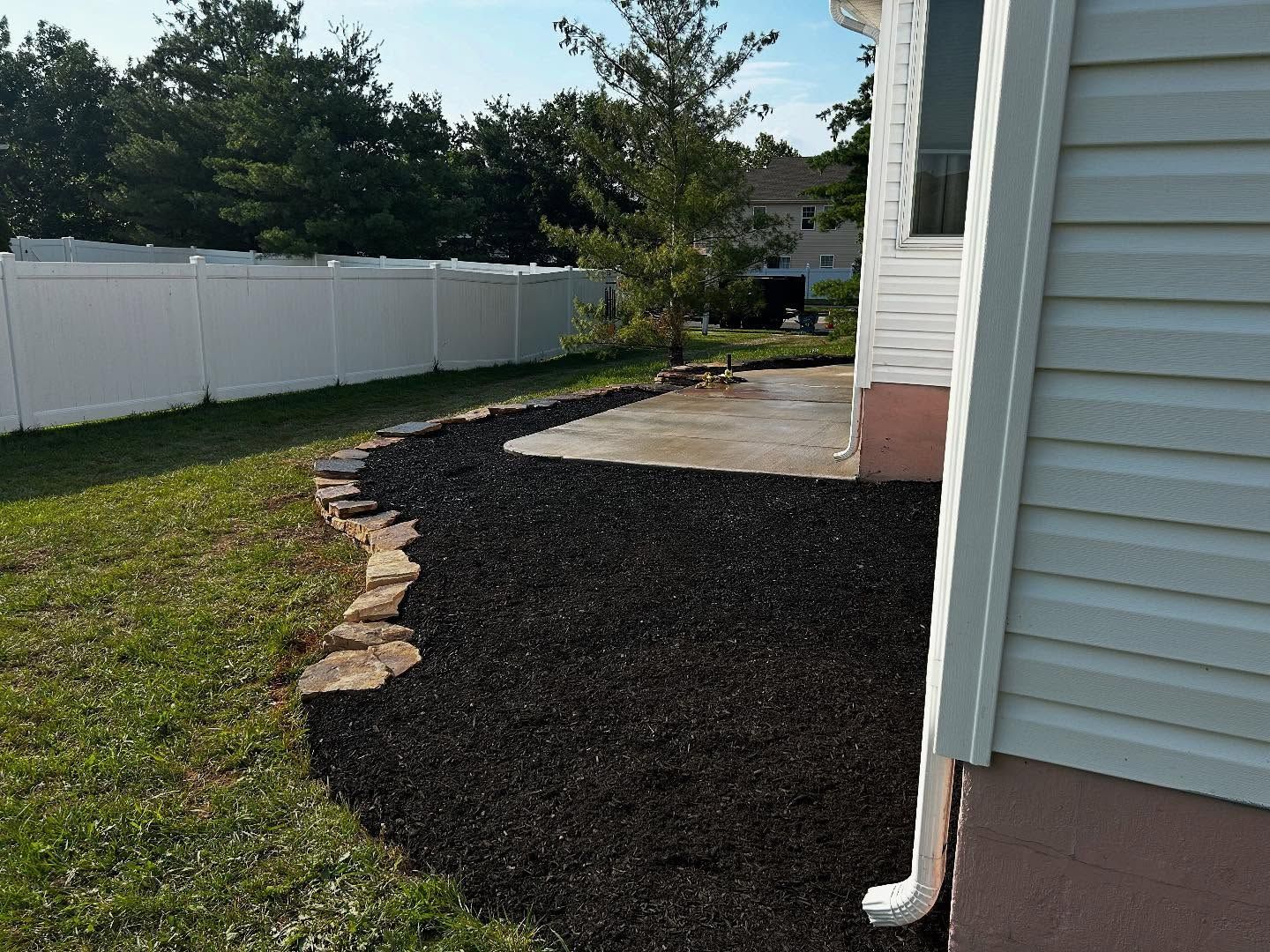 Backyard landscaping with black mulch, a concrete patio, and a white fence.