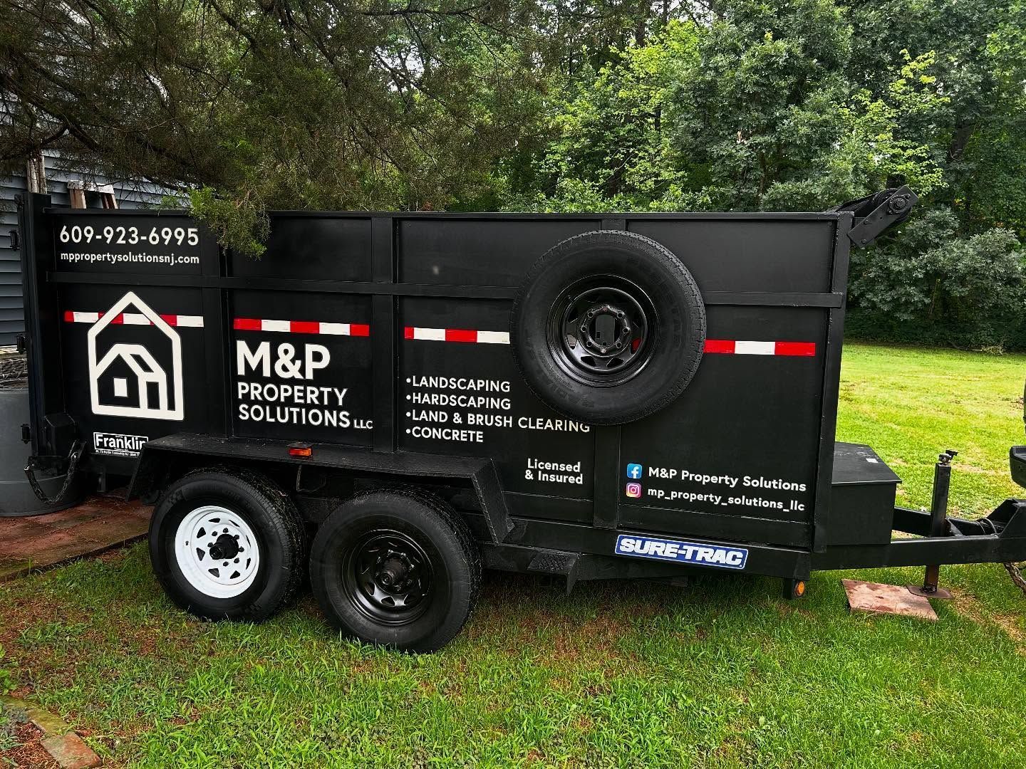 Black M&P Property Solutions dump trailer with spare tire. White/black logo, red accents on a lawn.