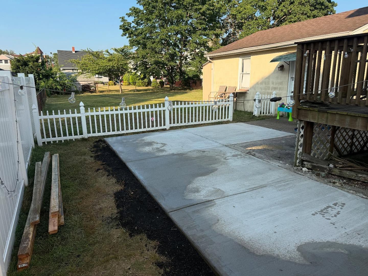 Concrete patio next to a wooden deck and a white picket fence, with grass in the background.