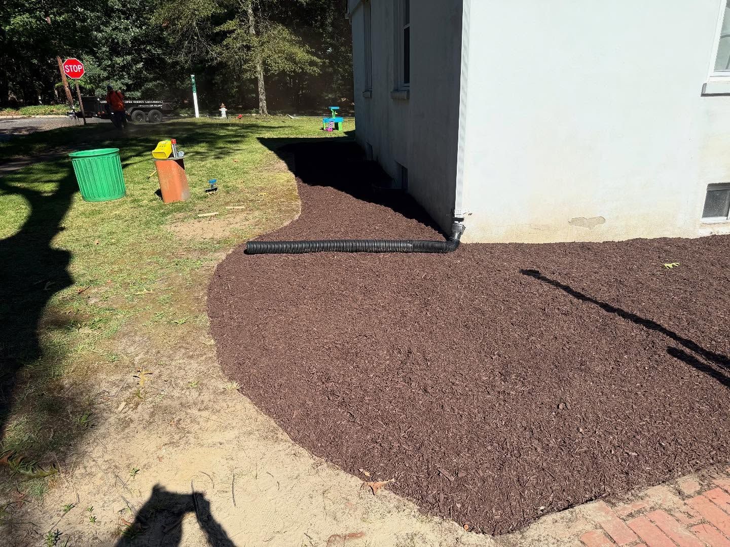 Brown mulch bed borders a white building, next to a grassy lawn with green and orange items.