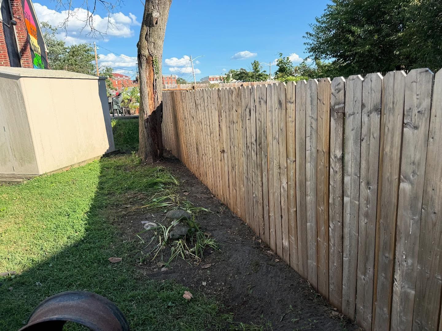 Wooden fence bordering a grassy yard with a tree and a concrete structure under a blue sky.