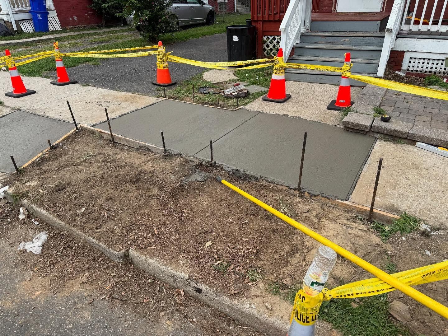 Freshly poured concrete sidewalk section, cordoned off with caution tape and cones.
