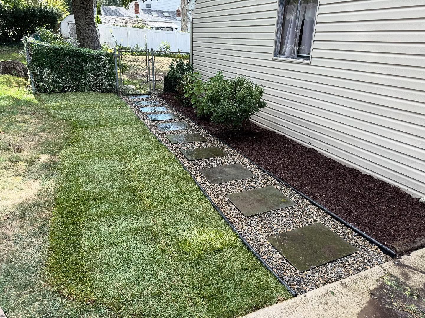 Path of stone pavers, surrounded by gravel and fresh lawn, next to a house with beige siding.