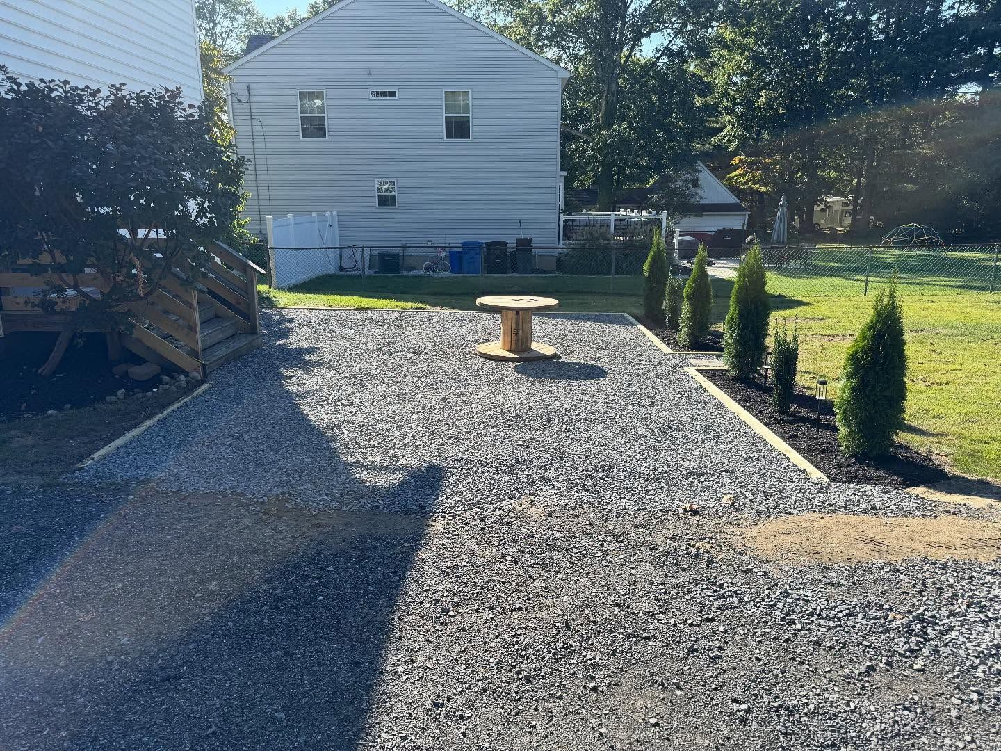 Gravel driveway leading to a light blue house. A wooden spool sits in the center. Green shrubs line the edge.