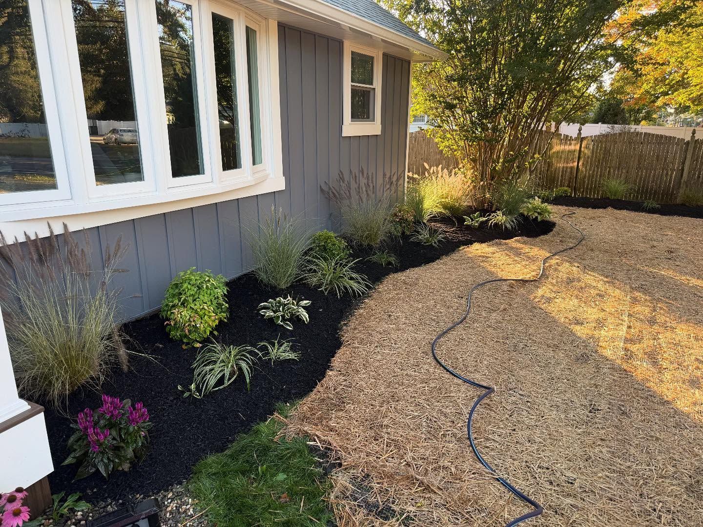 Landscaped house exterior with flower beds and a winding walkway. Dark mulch, gray siding, and a bay window.
