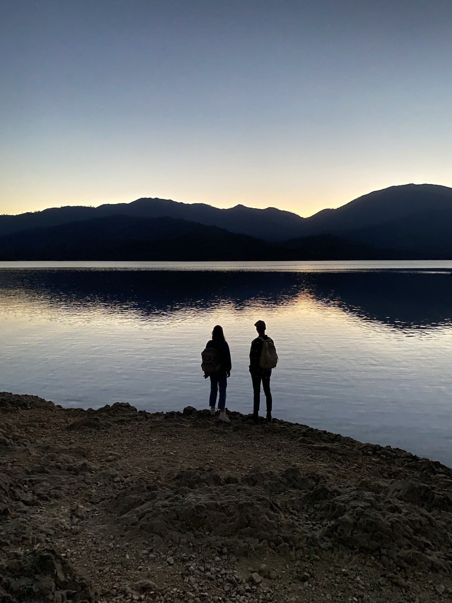 A couple stand on the edge of a lake surrounded by mountains during sunset as they talk about Northern California camping.