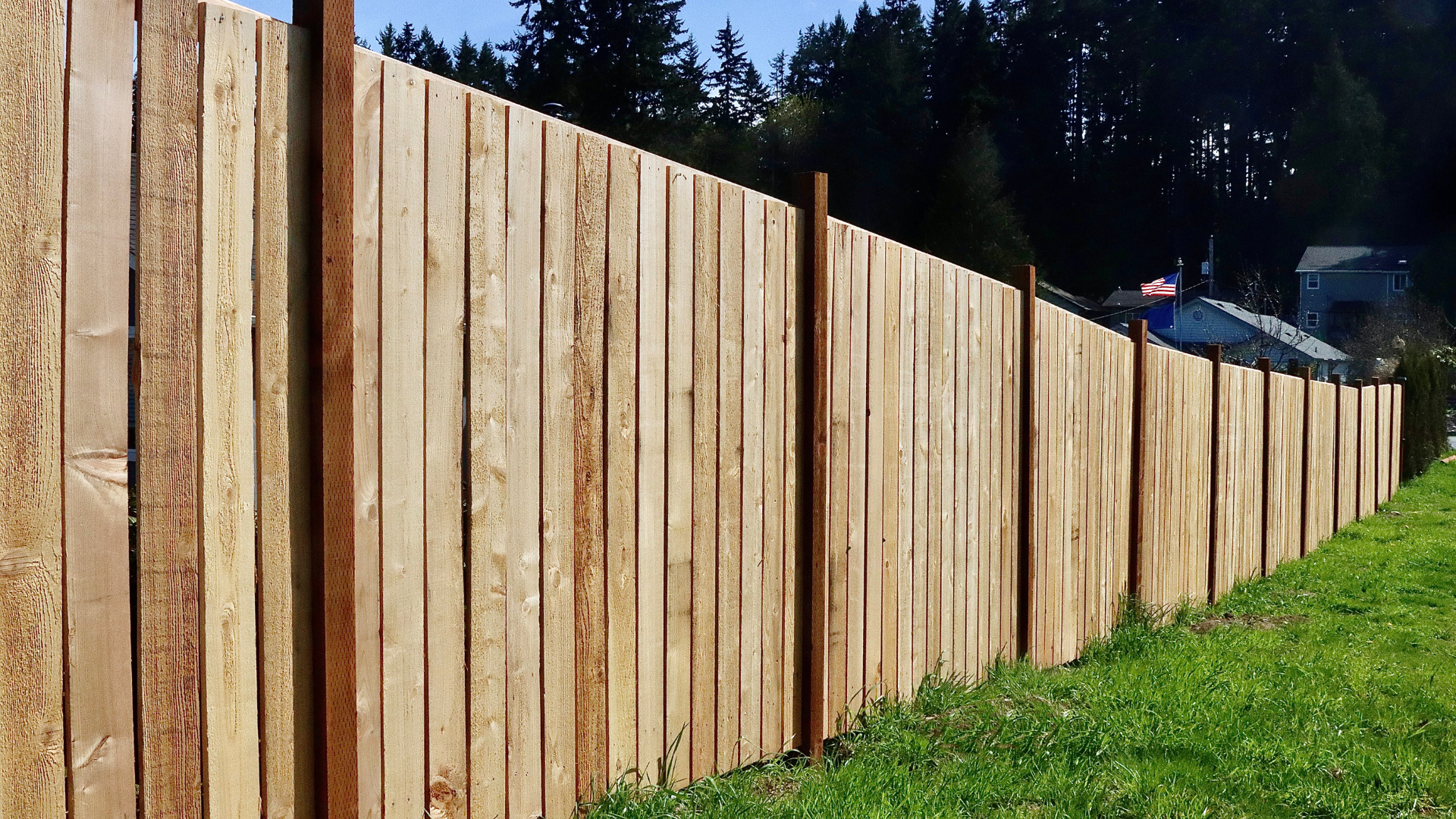 Wooden fence in a grassy yard, brown boards, tall posts, with trees and a building in the background.