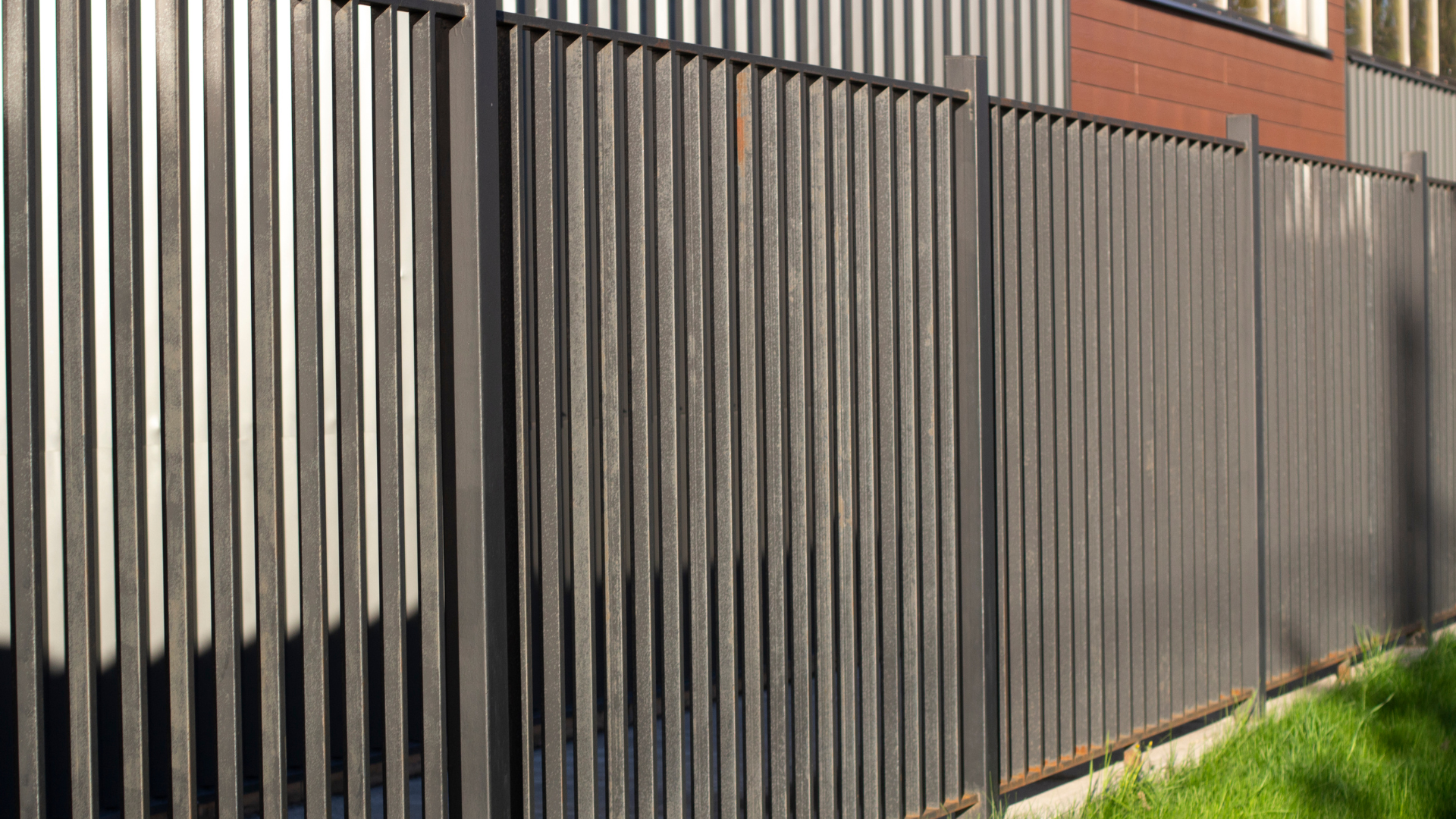 A tall, grey metal slat fence stands outdoors next to a patch of green grass and a modern building wall.