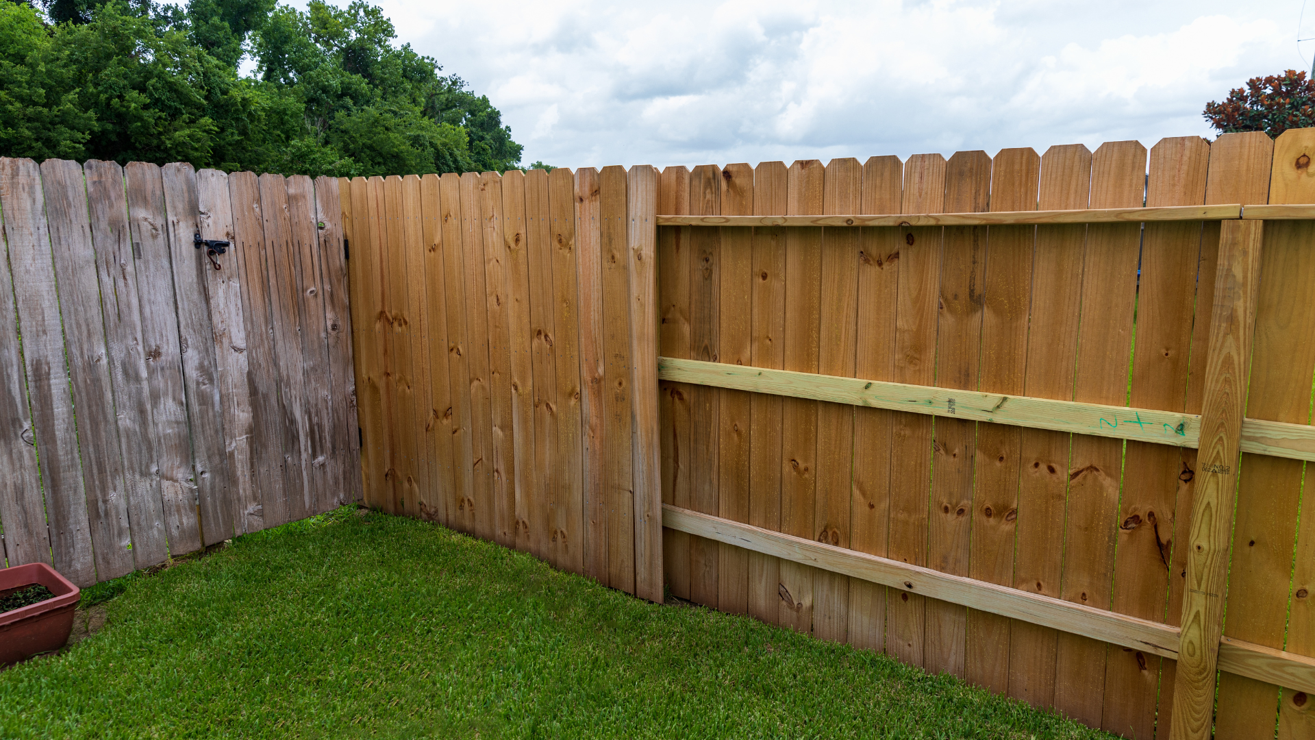 Wooden fence with a section of new boards and a section with weathered wood, grass in the foreground, cloudy sky.