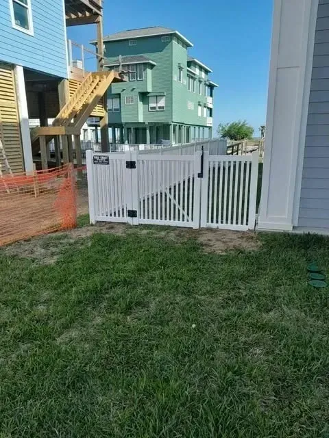White picket fence with gate, enclosing grassy yard, beach houses in the background.