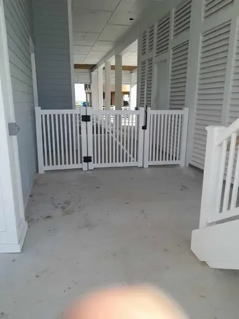 White picket fence with gate on a concrete porch, beside a house with shutters.