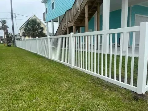White picket fence along a grassy lawn, in front of a turquoise house on stilts.