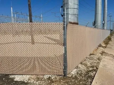 Tan privacy slats in a chain-link fence bordering a paved area, with power poles and a substation in the background.
