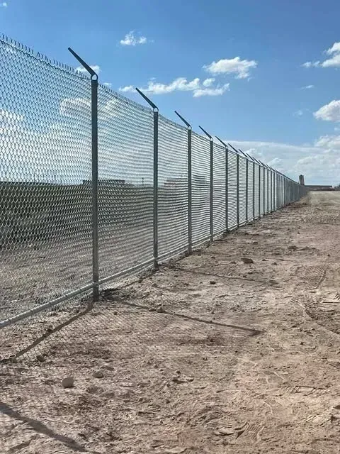 Chain-link fence with barbed wire on top, erected in a dirt field under a blue sky.