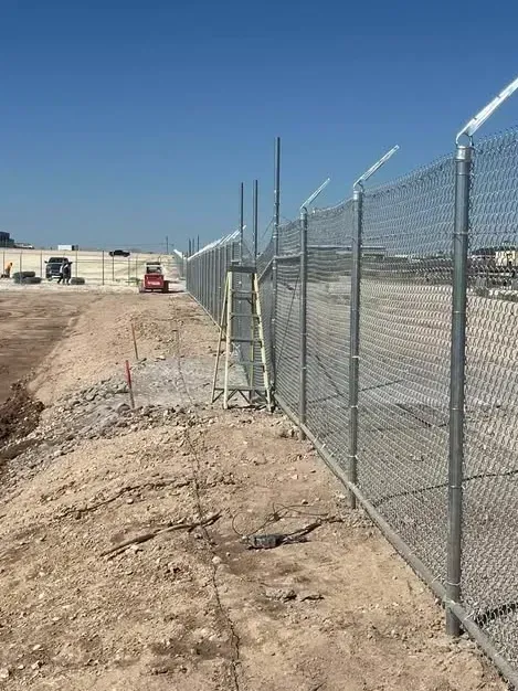 Chain-link fence topped with barbed wire runs along a dirt embankment, with construction equipment visible in the distance.