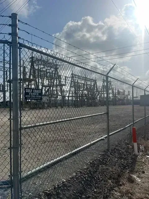 Chain-link fence with barbed wire surrounds an electrical substation on a sunny day.