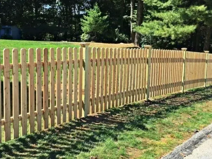 Wooden picket fence bordering a grassy yard, with trees in the background under a sunny sky.