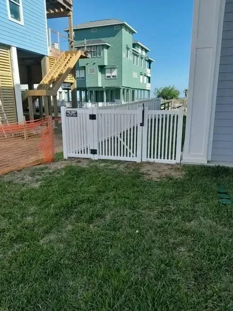 White picket fence and gate in front of a blue and green beach house, with grassy yard and stairs.